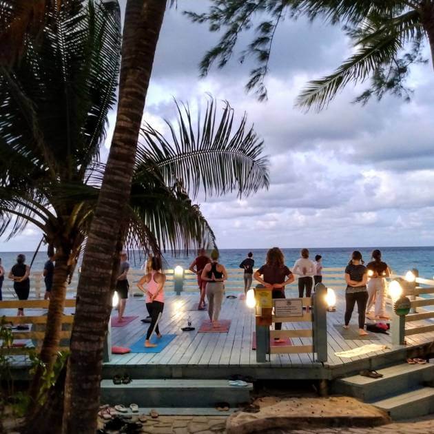Load image into Gallery viewer, Gathered group of yoga practitioners enjoying a session by the beach front Back to your Roots at Nevis with Amanda Parr - VendorYoga &amp; Scuba Diving - RETREAT - Zhoola
