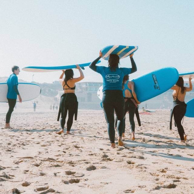 Girl gang carrying surfboards, walking beachside.Surf & Underwater Training with Nette, Augusto & Erika (Women Only) - Surfing & Yoga - Retreat - Zhoola